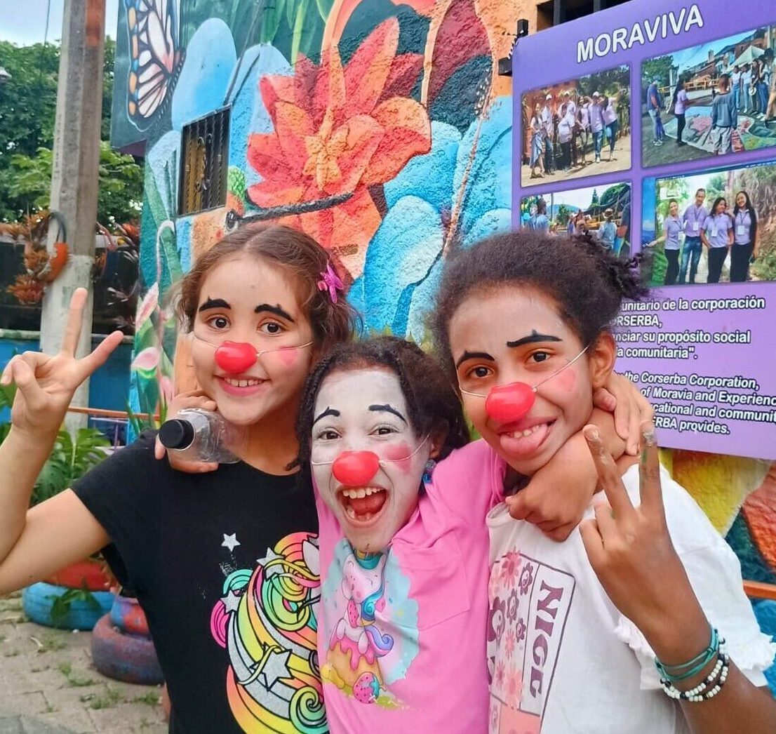 Tres niñas sonriendo y abrazadas, con narices de payaso, frente al mural de Moraviva en Moravia, Medellín